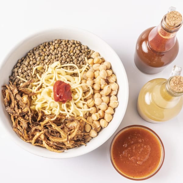 A bowl of Koshary with lentils, chickpeas, pasta, fried onions, and tomato sauce on a white background.