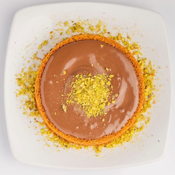 Middle Eastern kunafa dessert with chocolate topping and crushed pistachios on a white plate.