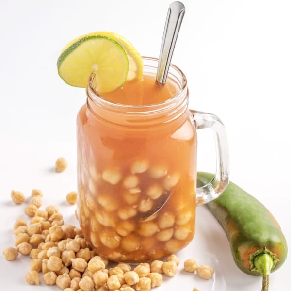 A glass mug of Middle-Eastern hummus soup with chickpeas, lime, and a green chili on a white surface.