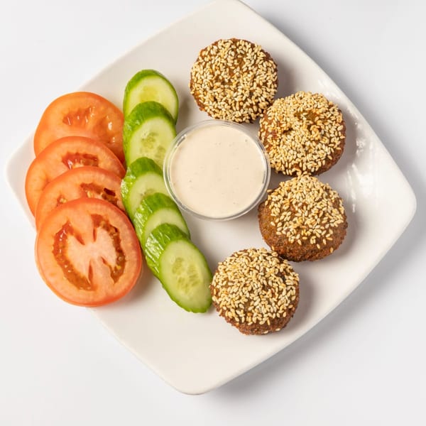 Falafel with sesame seeds, sliced tomatoes, cucumbers, and a dipping sauce on a white plate.