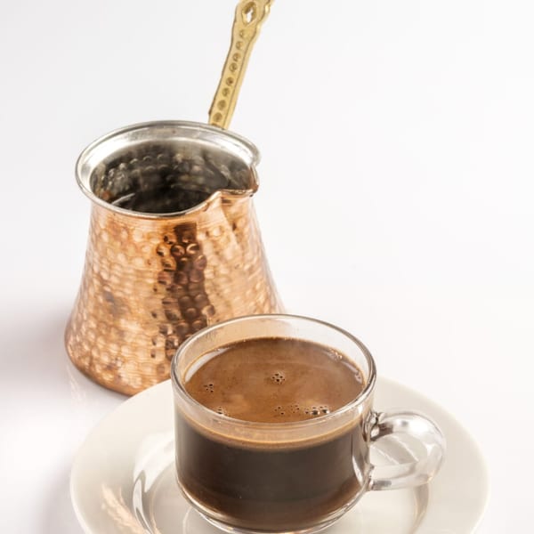 A clear glass cup of dark, rich Middle Eastern coffee next to a traditional hammered copper cezve.